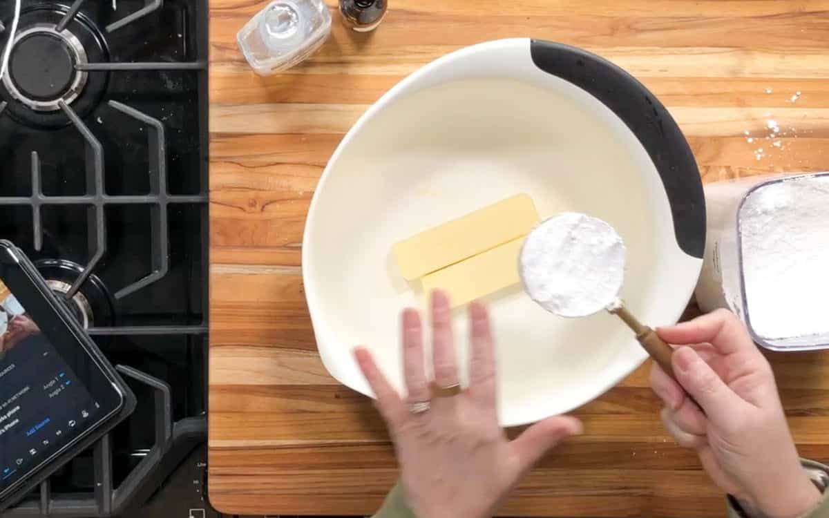 A person holds a measuring cup of powdered sugar above a large mixing bowl containing two sticks of butter on a wooden countertop. A tablet, box of flour, and small container are nearby.