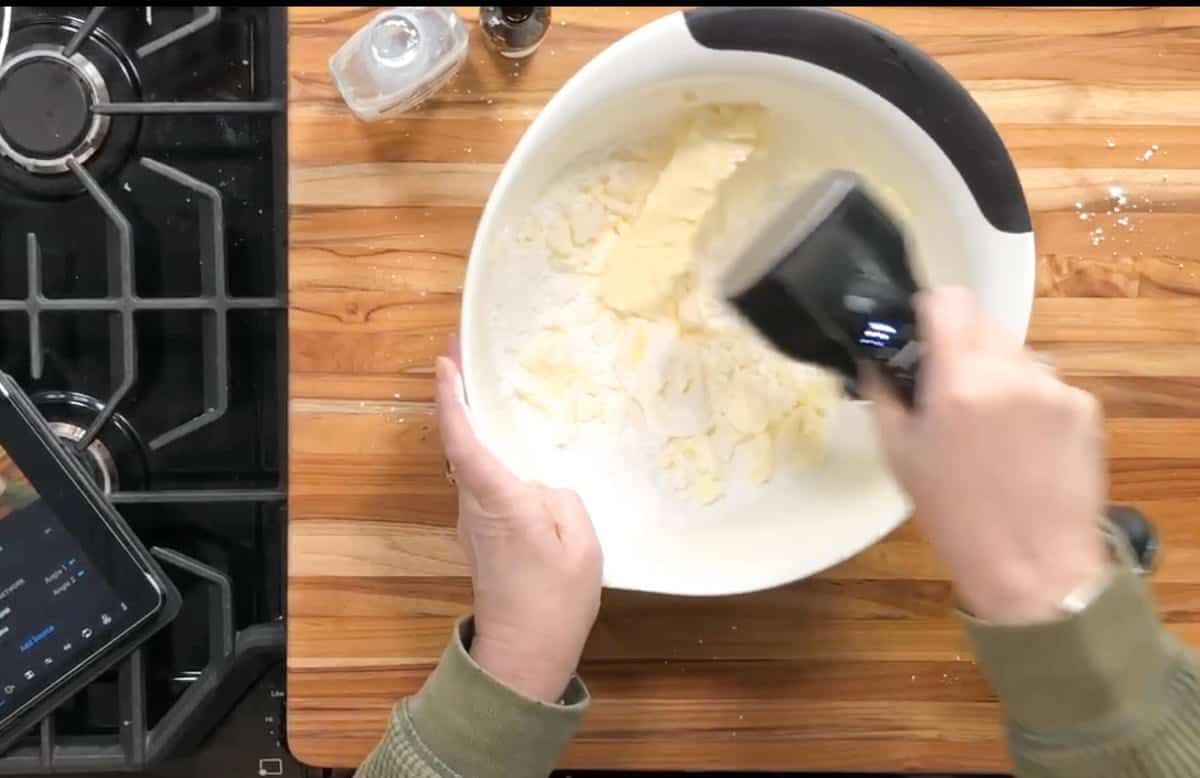 A person mixes butter and flour in a white bowl with a black hand mixer on a wooden countertop next to a stove, salt shaker, and a tablet.