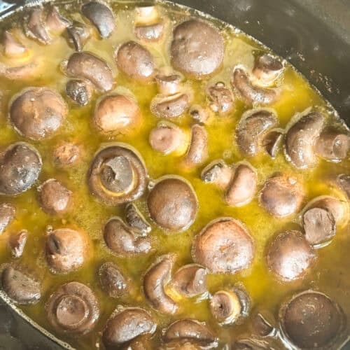 A close-up view of whole mushrooms simmering in a yellowish liquid, possibly broth or butter, inside a slow cooker or pot.