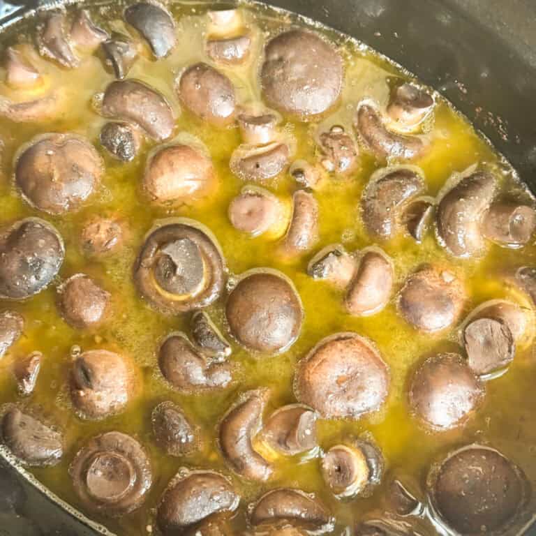 A close-up view of whole mushrooms simmering in a yellowish liquid, possibly broth or butter, inside a slow cooker or pot.