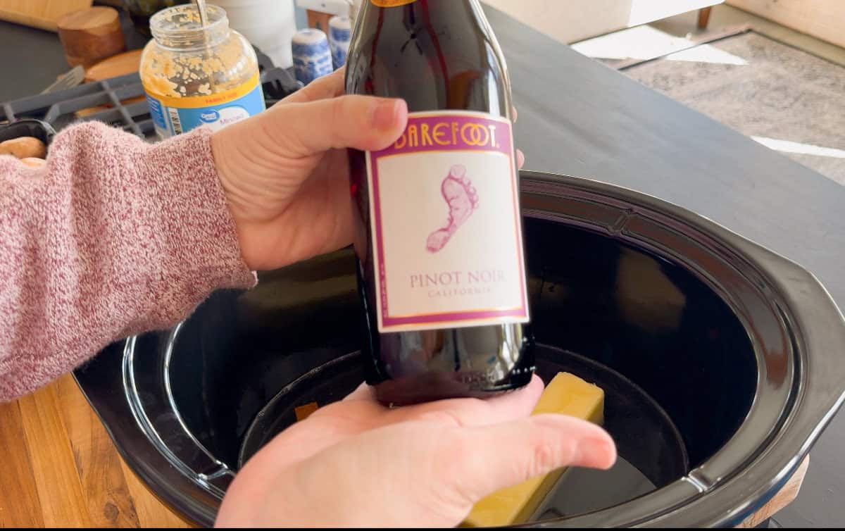 A person holds a bottle of Barefoot Pinot Noir wine over an oval-shaped black slow cooker with two sticks of butter inside. A jar of minced garlic and other kitchen items are visible in the background.