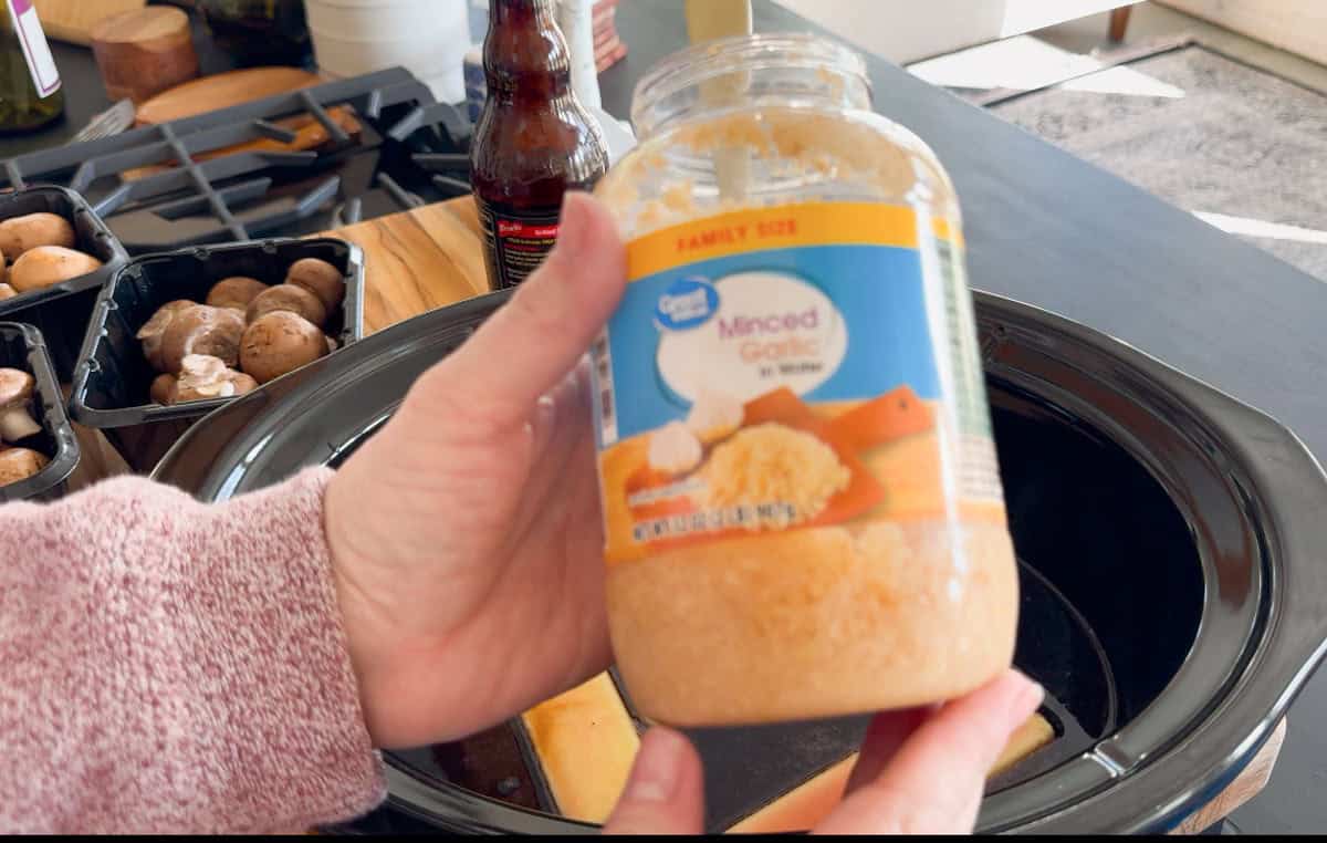 A person holds a large plastic jar of minced garlic over a slow cooker, with packages of mushrooms and kitchen items visible in the background.