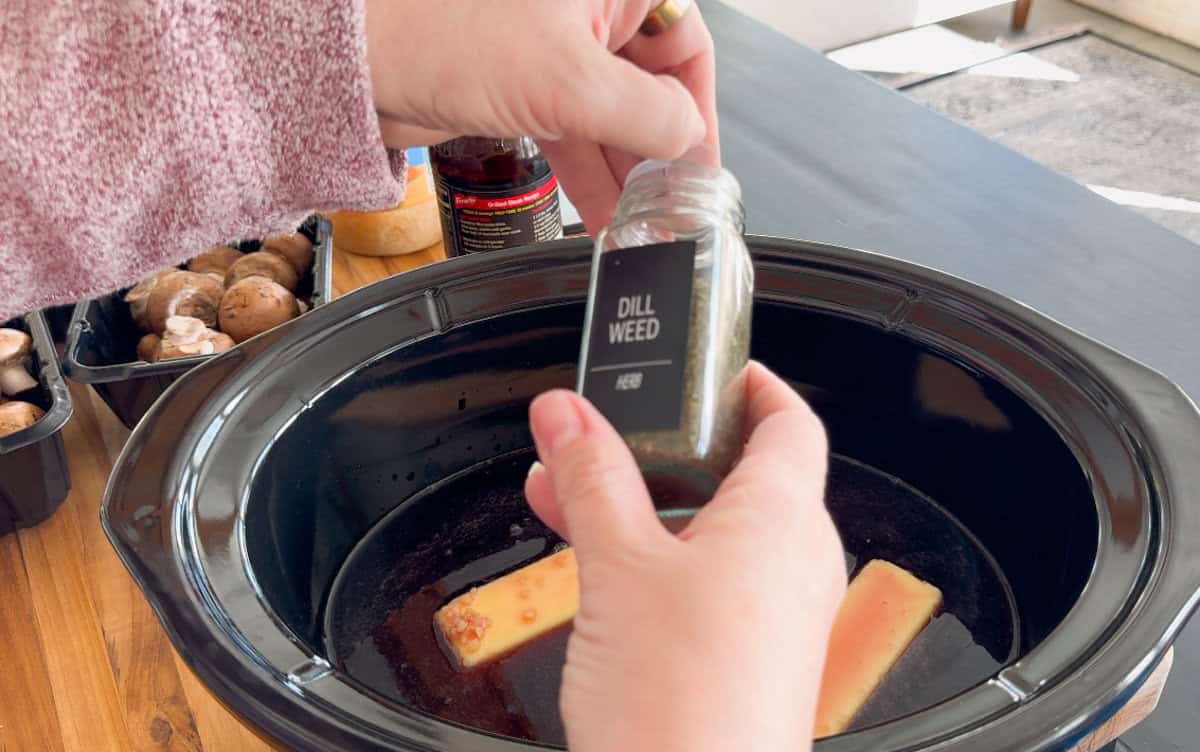 A person sprinkles dill weed from a jar into a slow cooker containing two butter sticks. Nearby, mushrooms and seasonings are visible on a wooden table.