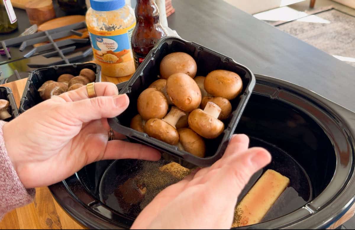 A person holds a black plastic container of brown mushrooms over a slow cooker. Inside the slow cooker are a block of butter and seasoning. A jar of peanut butter and other ingredients are visible on the counter.