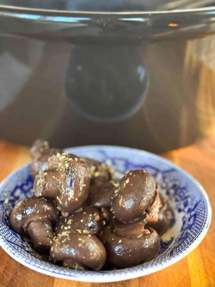A blue and white patterned bowl filled with cooked mushrooms topped with herbs sits on a wooden surface in front of a black slow cooker.