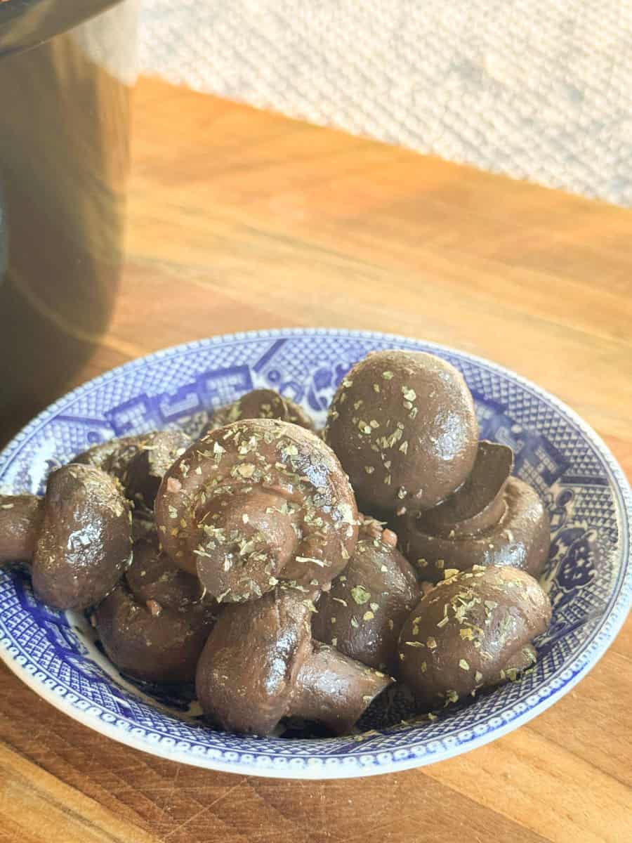 A blue and white patterned bowl filled with seasoned, cooked whole mushrooms sits on a wooden surface with a textured white background.