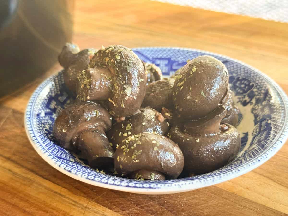 A blue and white patterned bowl filled with cooked mushrooms seasoned with herbs sits on a wooden surface.