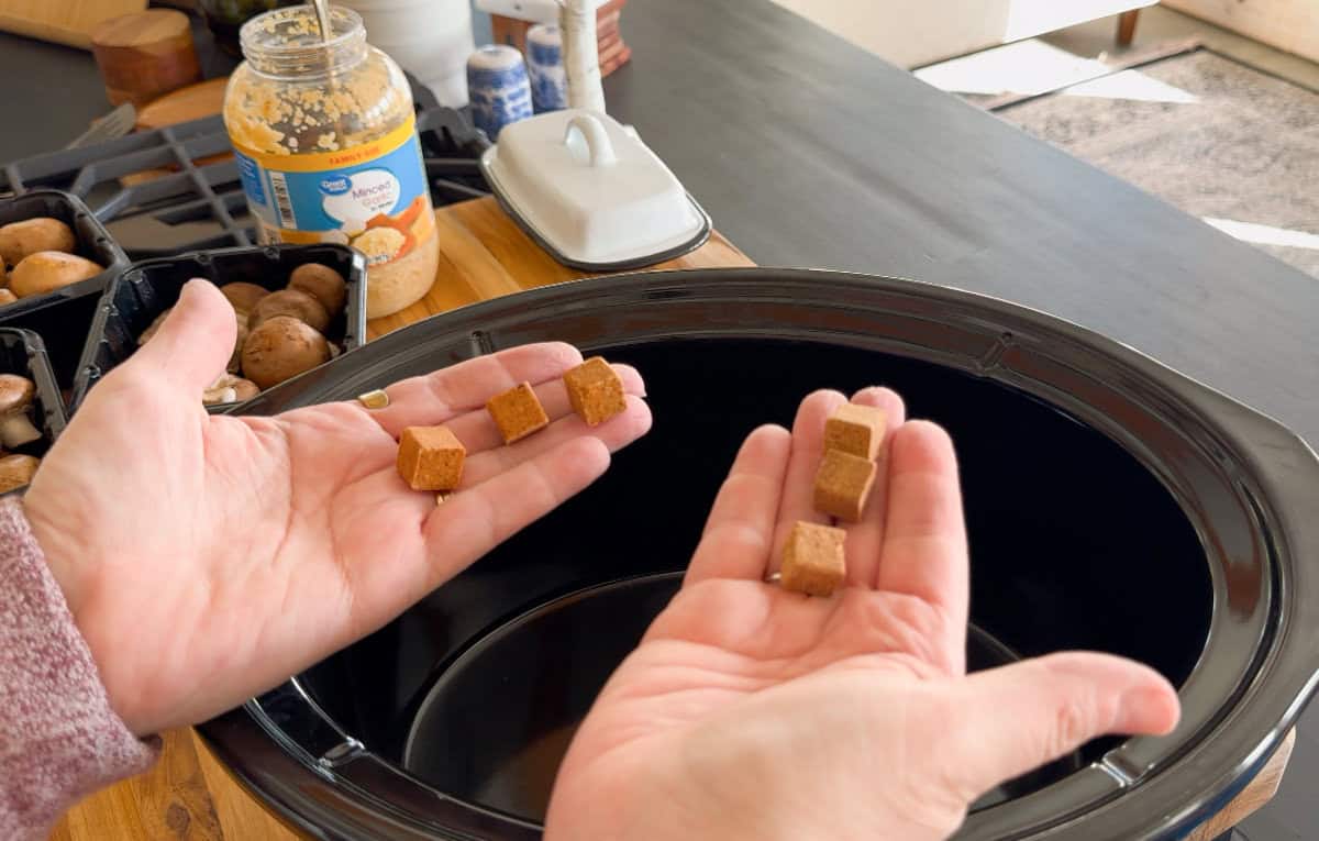 A person holds cube-shaped food pieces over an empty black slow cooker, with a jar of minced garlic, mushrooms, and other kitchen items visible on the counter nearby.