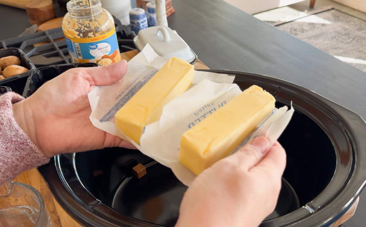 Two hands hold two sticks of butter over an open slow cooker, with a jar of minced garlic and various kitchen items visible in the background.