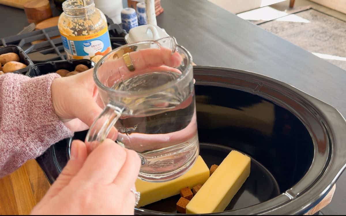 A person holds a glass measuring cup filled with water above a slow cooker containing two sticks of butter and two brown bouillon cubes. The scene includes a jar of minced garlic and kitchen items in the background.