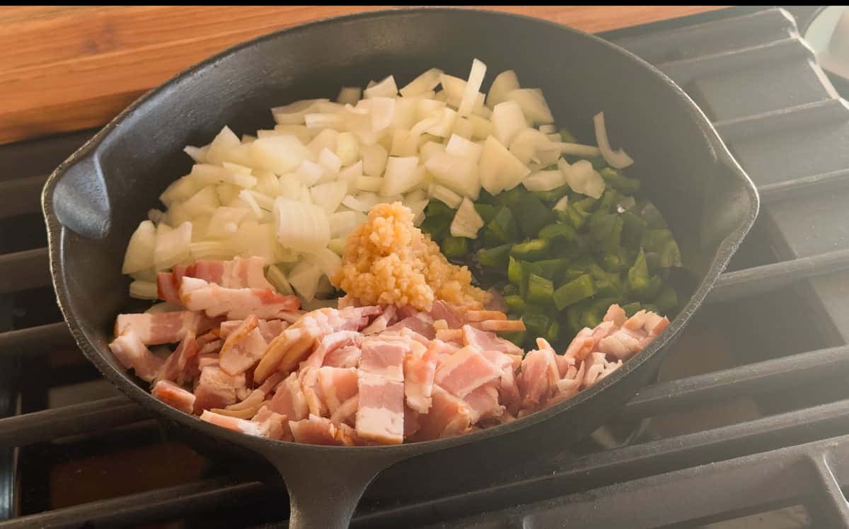 A cast iron skillet on a stove contains chopped onions, diced green peppers, minced garlic, and chopped bacon prepared for cooking.