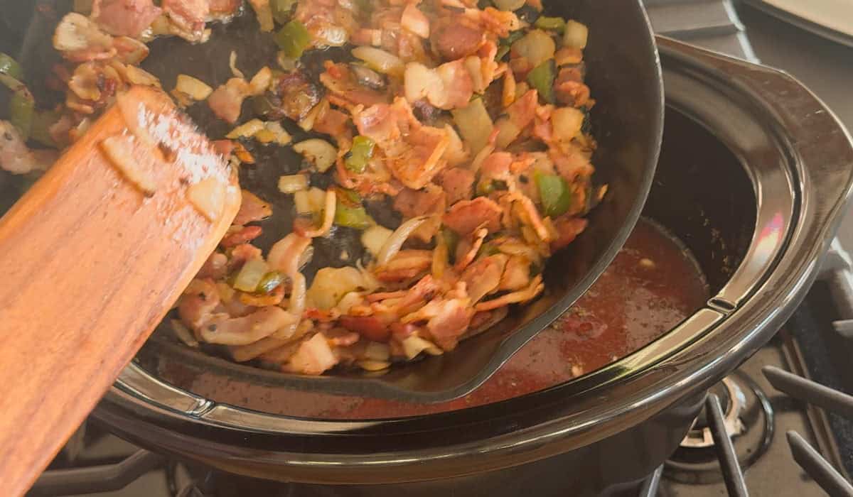 A person uses a wooden spatula to pour a cooked mixture of onions, green peppers, and bacon from a skillet into a slow cooker containing a red liquid.