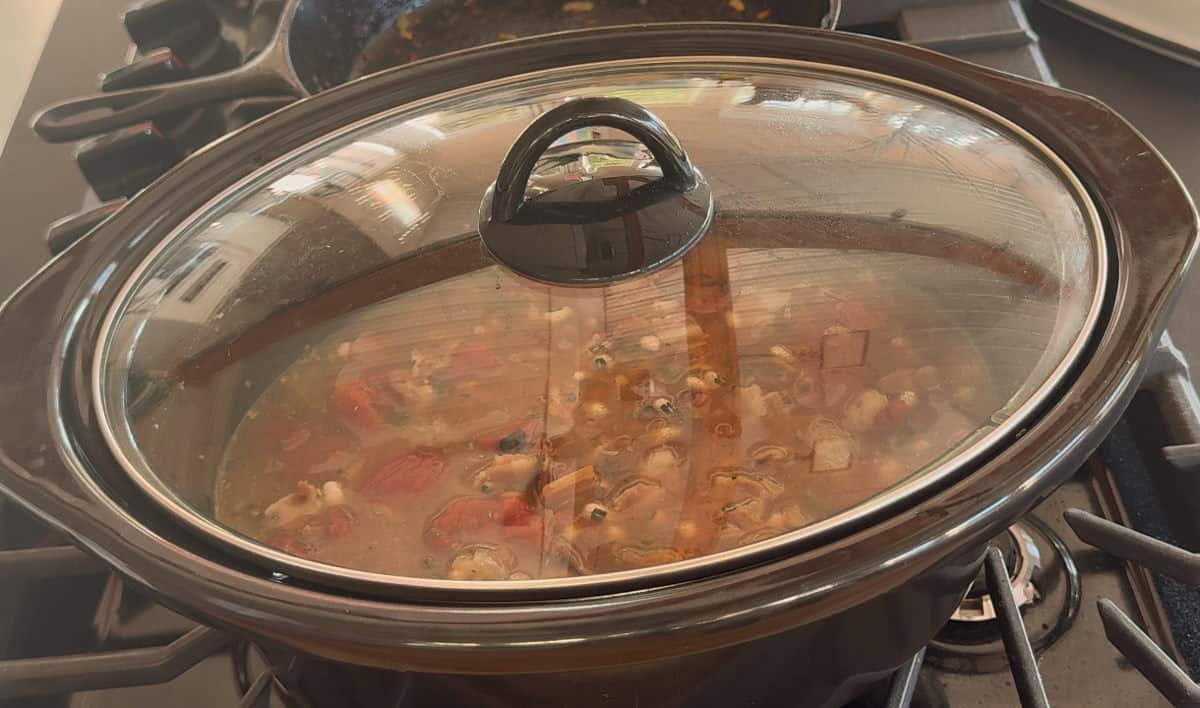 A slow cooker with a glass lid is simmering a stew that contains visible chunks of vegetables and beans. The slow cooker is placed on a stovetop.