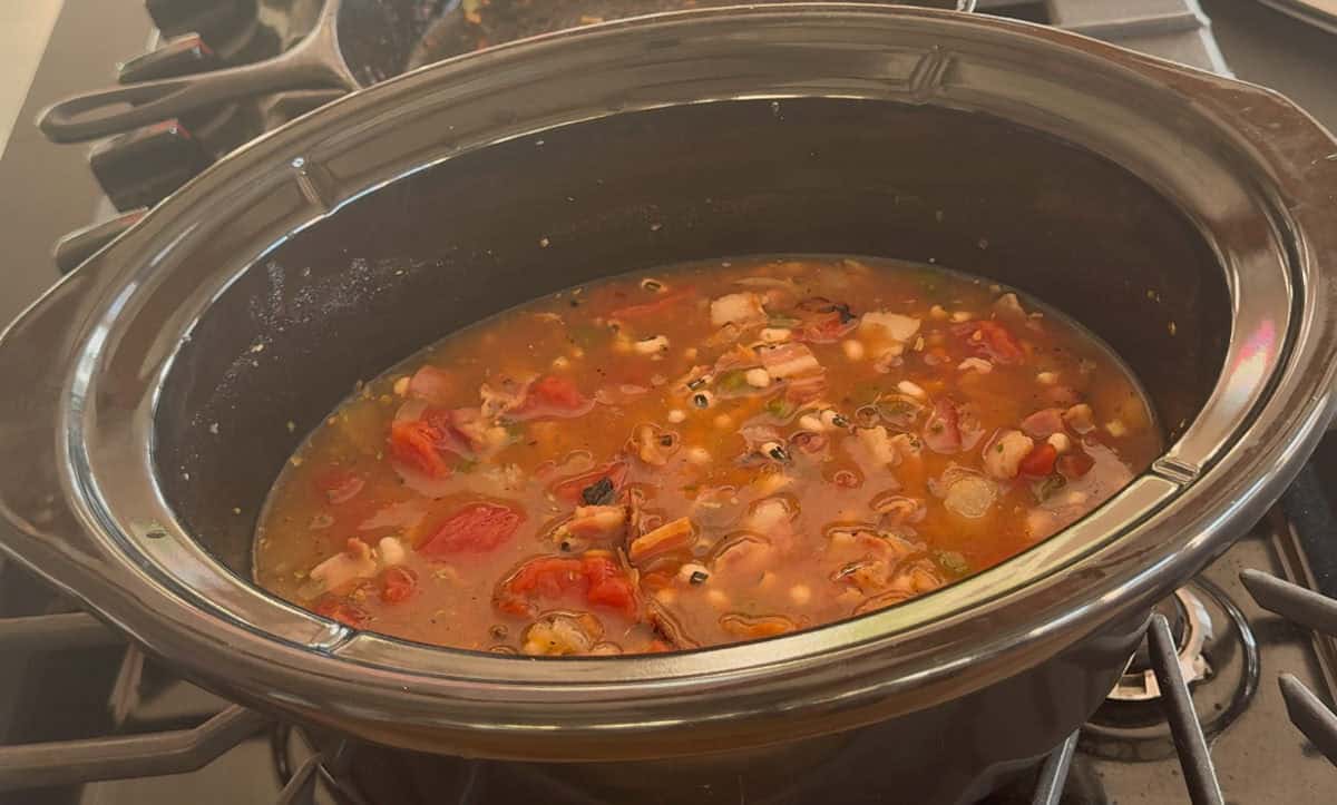 A slow cooker filled with a chunky soup or stew containing tomatoes, beans, and various vegetables, sits on a gas stove with burners visible in the background.