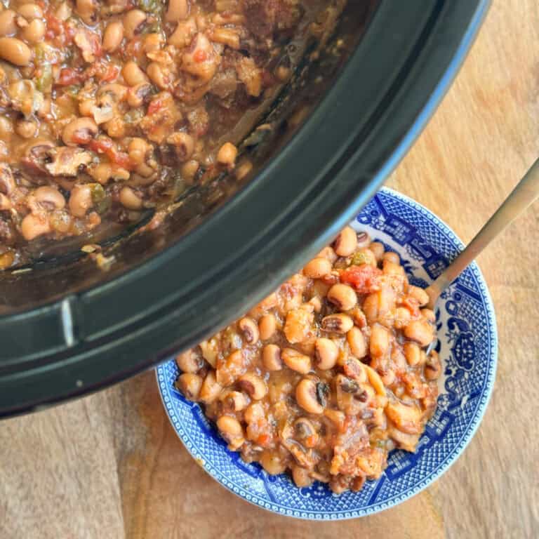 A close-up view of black-eyed peas with vegetables and sauce in a slow cooker, with a portion served in a blue and white patterned bowl on a wooden surface. A spoon is in the bowl.