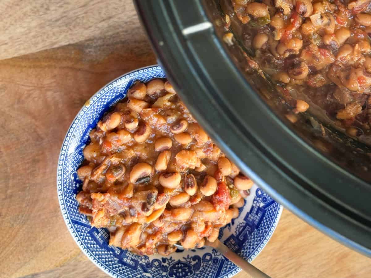 A bowl of cooked black-eyed peas with tomatoes and vegetables sits next to a slow cooker filled with more of the same dish, on a wooden surface. A spoon rests in the bowl.