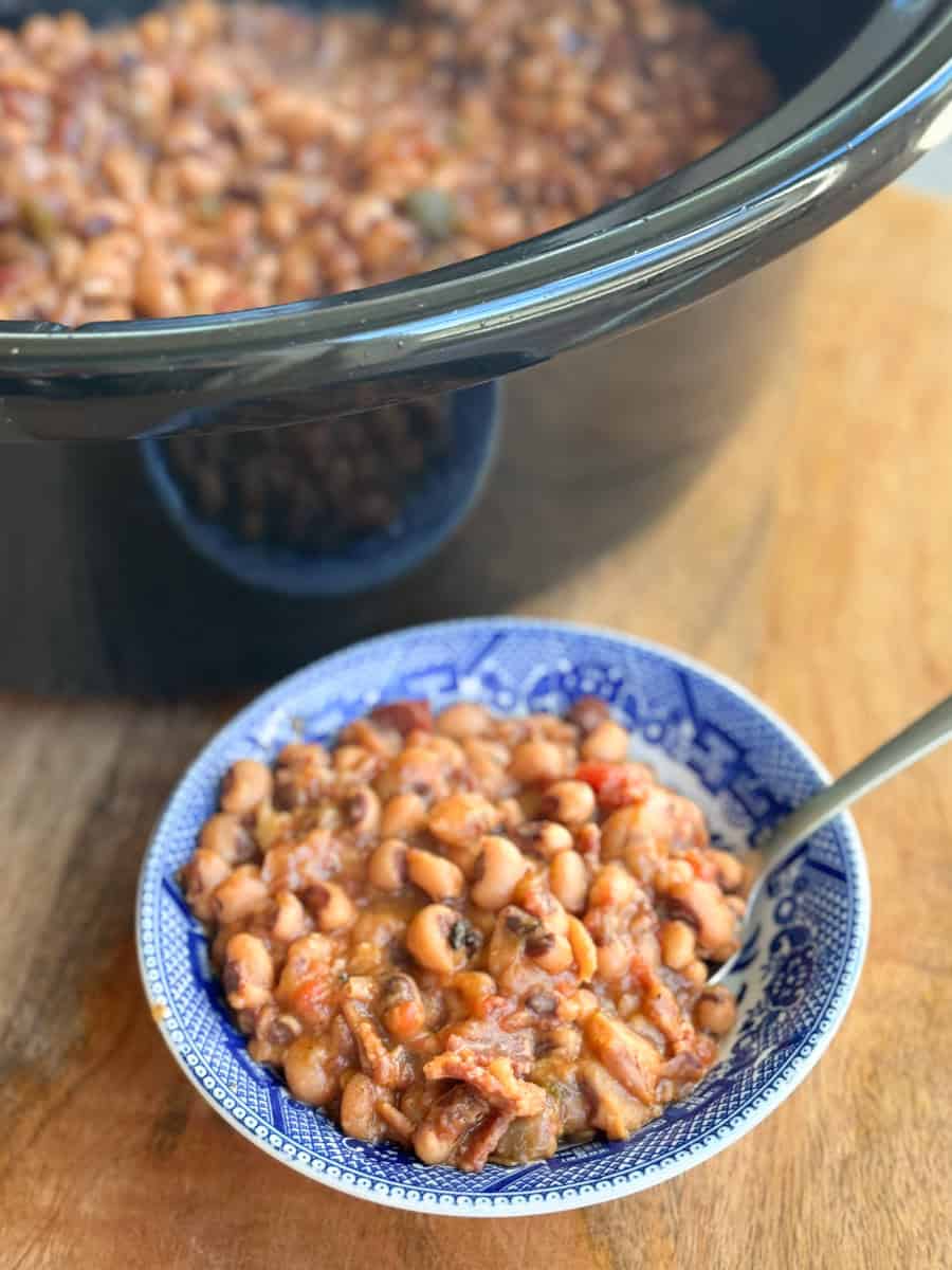 A blue and white bowl filled with a bean stew sits on a wooden surface in front of a black slow cooker that is also filled with the same stew. A spoon is placed in the bowl.
