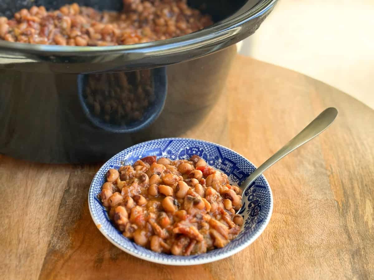 A blue and white patterned bowl filled with cooked beans in a tomato-based sauce, with a spoon resting in the bowl. In the background, a black slow cooker contains more of the same bean dish. The bowl sits on a wooden surface.