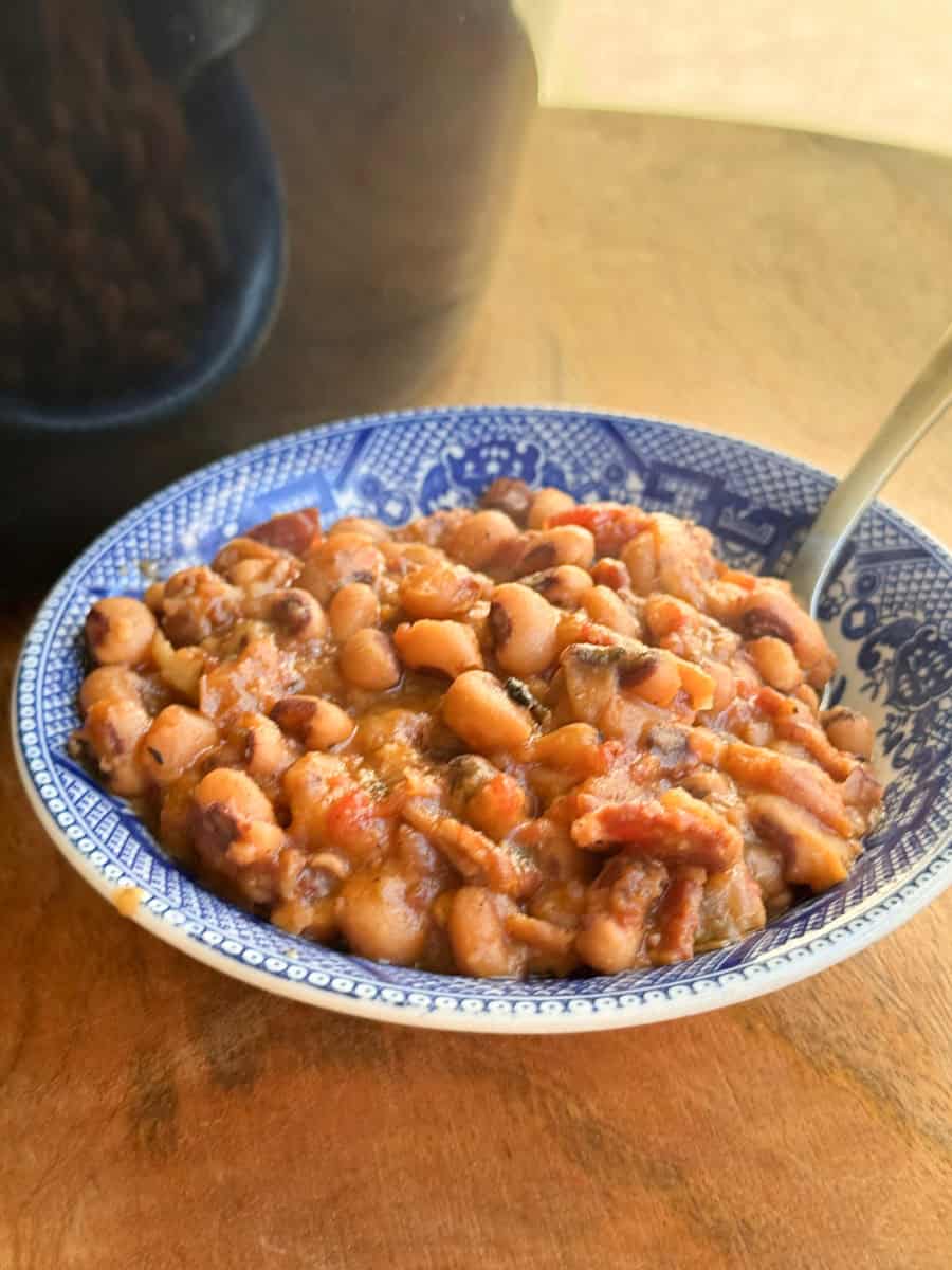 A blue and white patterned bowl filled with cooked black-eyed peas mixed with pieces of bacon and tomatoes, placed on a wooden surface with a spoon in the bowl.