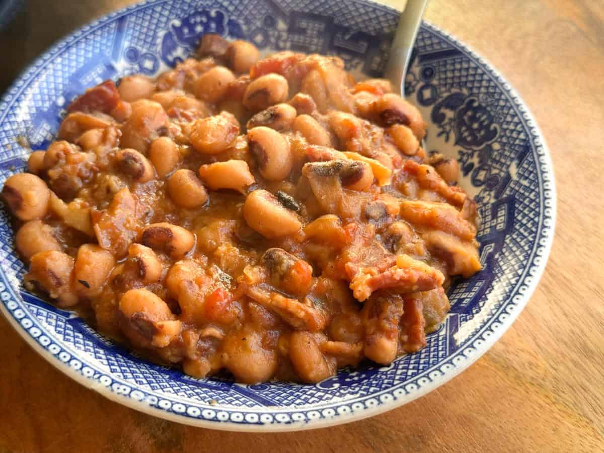 A blue and white patterned bowl filled with cooked black-eyed peas, visible tomato pieces, and sliced bacon, with a spoon resting in the bowl on a wooden surface.