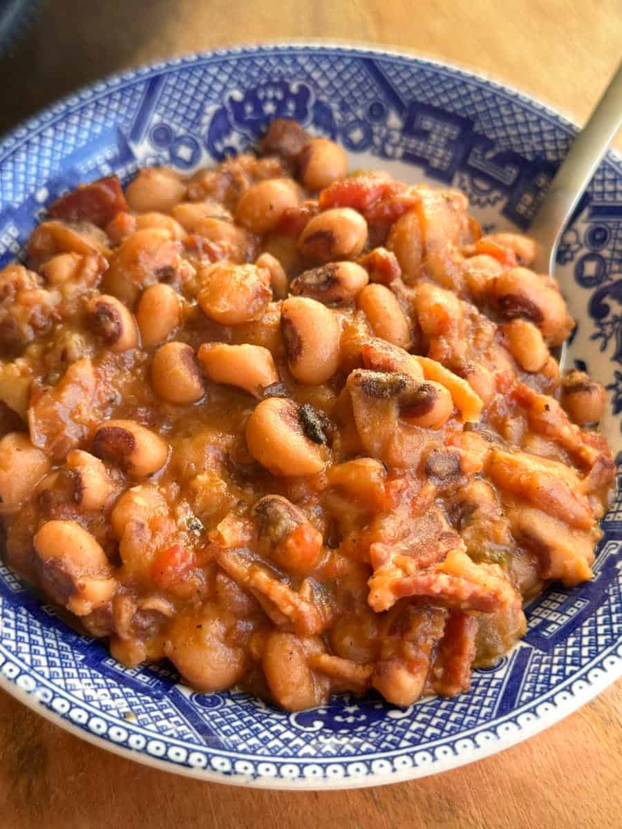 A close-up of a blue patterned bowl filled with cooked black-eyed peas, diced tomatoes, bacon, and vegetables, with a spoon resting inside the bowl.