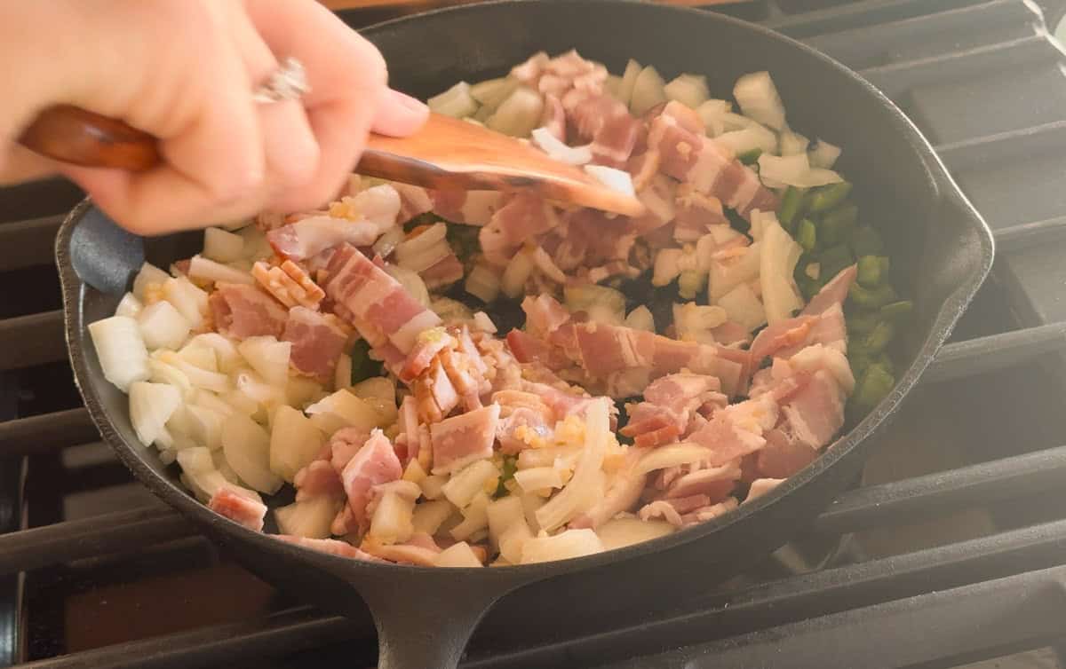 A hand stirs chopped bacon, onions, and bell peppers in a black cast iron skillet on a gas stove.