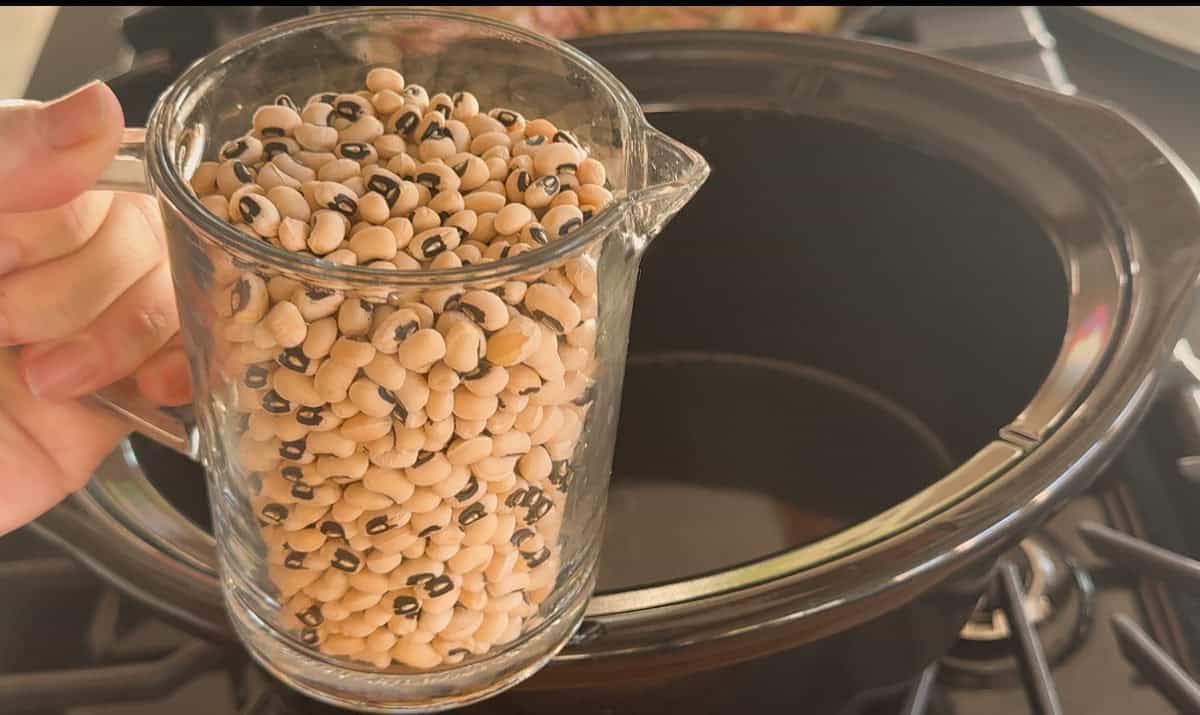 A hand holds a glass measuring cup filled with dry black-eyed peas above an empty black slow cooker on a stovetop.