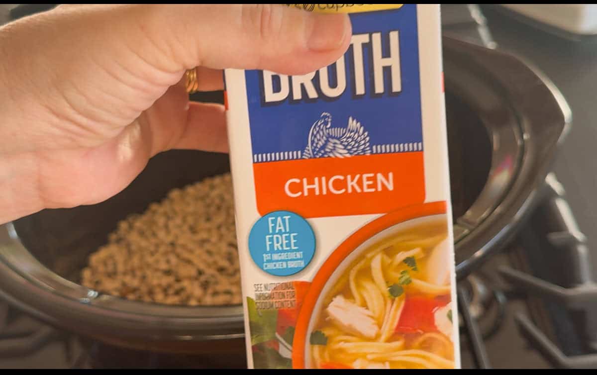 A hand holds a carton of fat-free chicken broth above a slow cooker filled with uncooked beans. The carton shows an image of chicken soup and text indicating it is chicken-flavored broth.