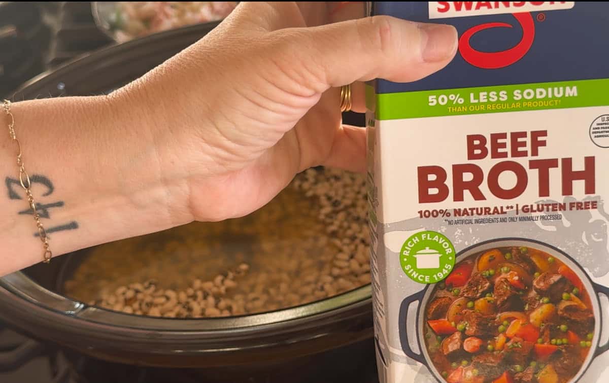 A hand holding a Swanson beef broth carton labeled “50% Less Sodium” and “Gluten Free” over a slow cooker filled with beans. A tattoo with numbers is visible on the person's wrist.