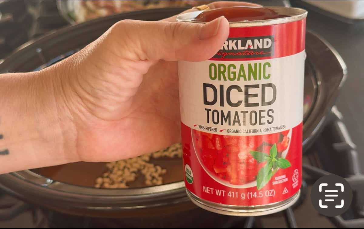 A hand holds a can of Kirkland Signature Organic Diced Tomatoes above an open slow cooker that contains beans. The can is 411 grams (14.5 oz) and features an image of diced tomatoes and basil.
