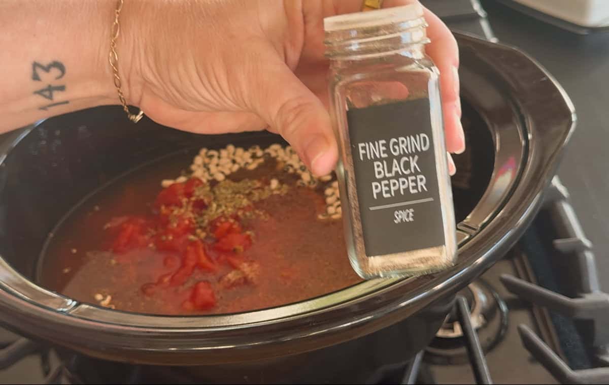 A hand holding a jar labeled "Fine Grind Black Pepper" above a slow cooker containing liquid, diced tomatoes, herbs, and grains on a stovetop.