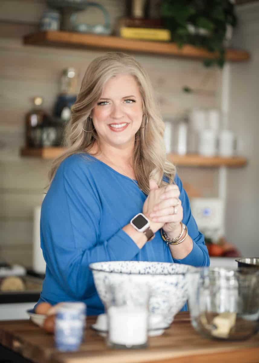 A woman with long blonde hair, wearing a blue top and a smartwatch, stands in a kitchen in front of a wooden counter with mixing bowls, a cup, an egg, and a glass container. She is smiling and clasping her hands.