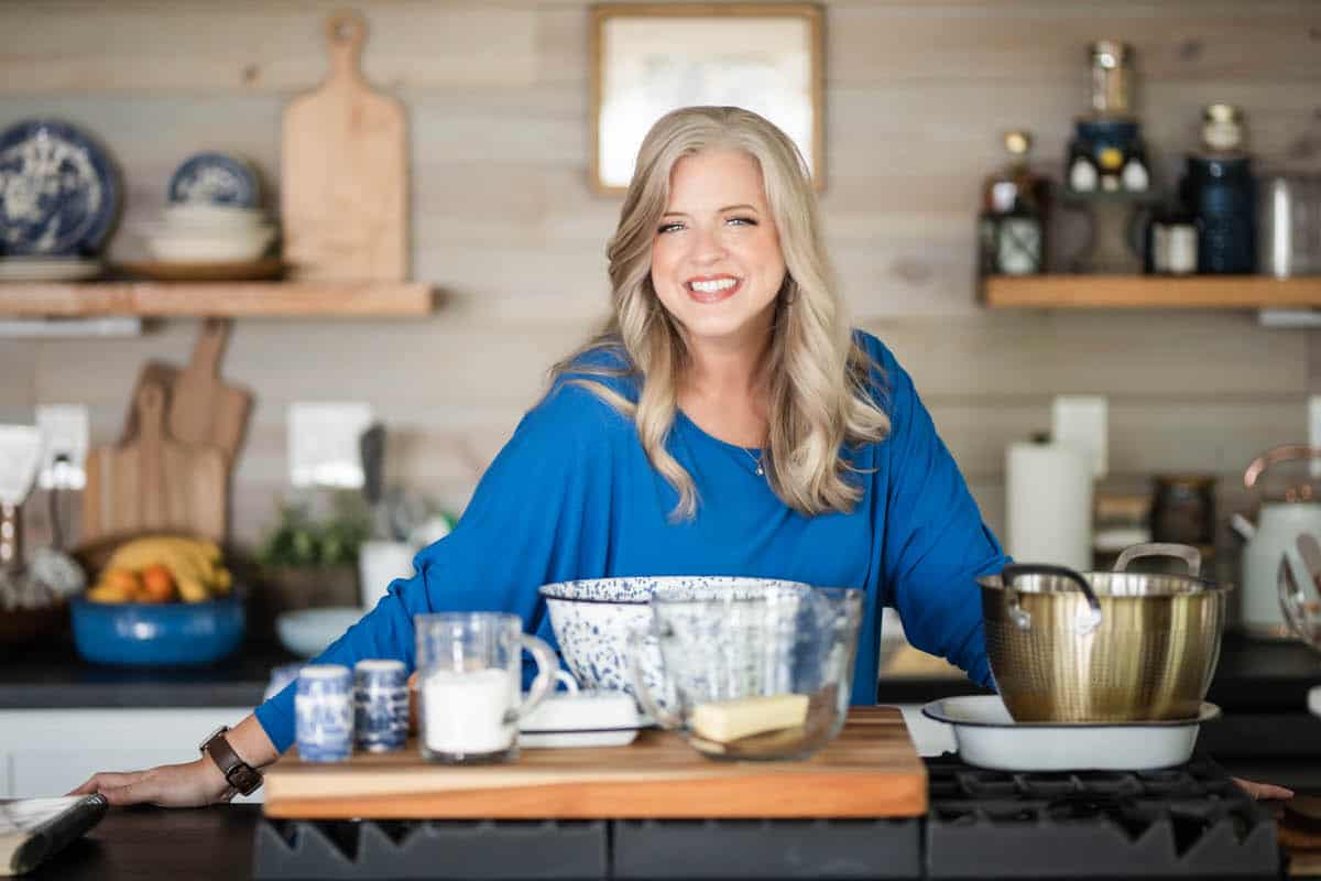A woman with long blonde hair, wearing a bright blue top, stands smiling in a modern kitchen behind a counter with mixing bowls, measuring cups, a stick of butter, and kitchen utensils. Shelves with dishes and decor are in the background.