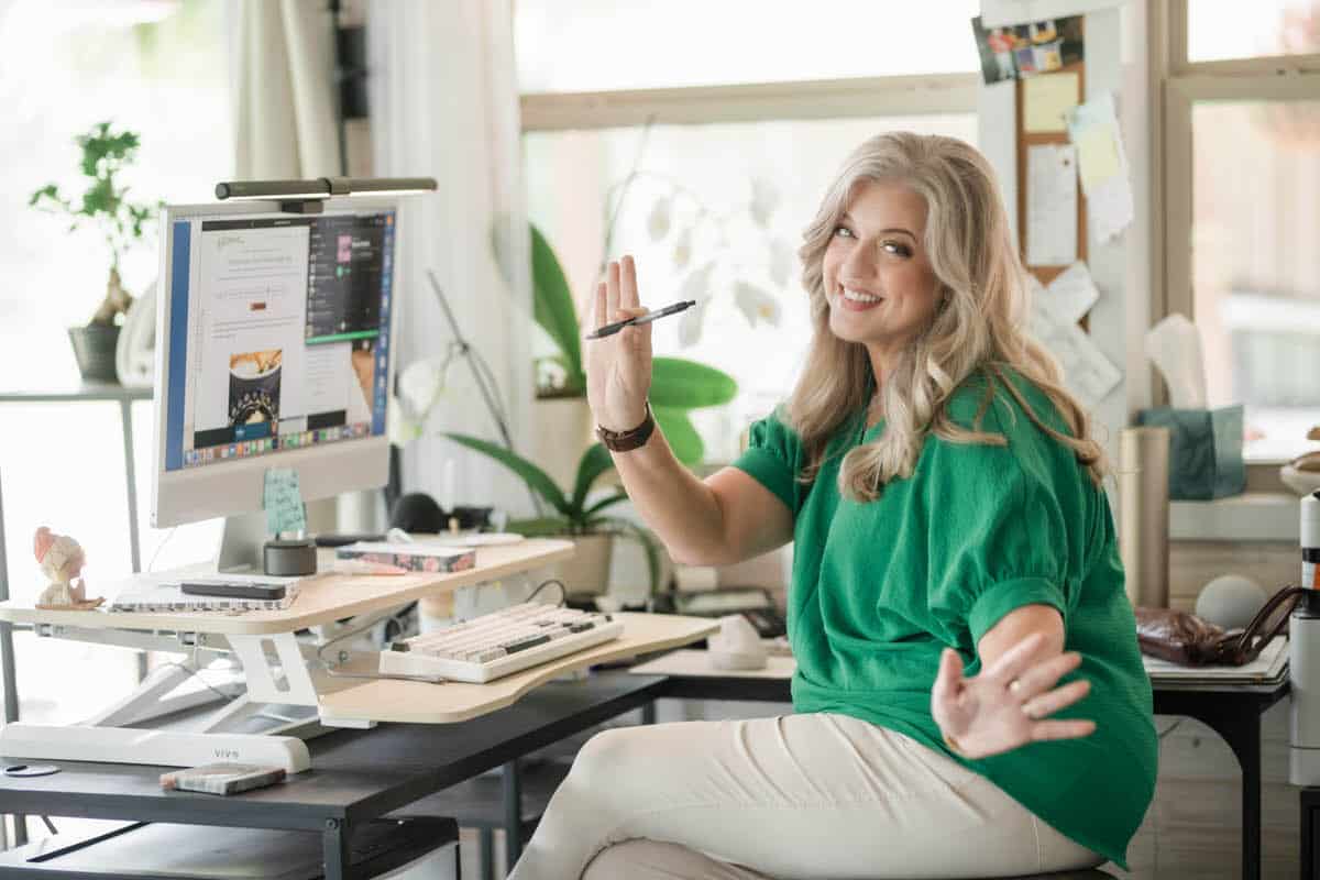 A woman with long, blonde hair wearing a green top sits at a desk with a computer, smiling and holding a pen in one hand while gesturing with the other; the workspace appears organized and well-lit.