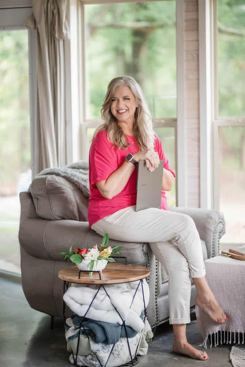 A woman with long blonde hair wearing a bright pink top and light pants sits on a gray couch, holding a tablet. She is barefoot and smiling, with a basket and small table with flowers beside her. Large windows are in the background.