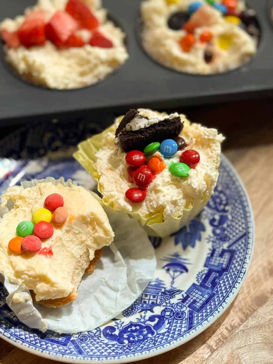 A plate with two cupcakes topped with candy and an Oreo cookie, one partially unwrapped and bitten. In the background, a muffin tin holds more cupcakes with various toppings.
