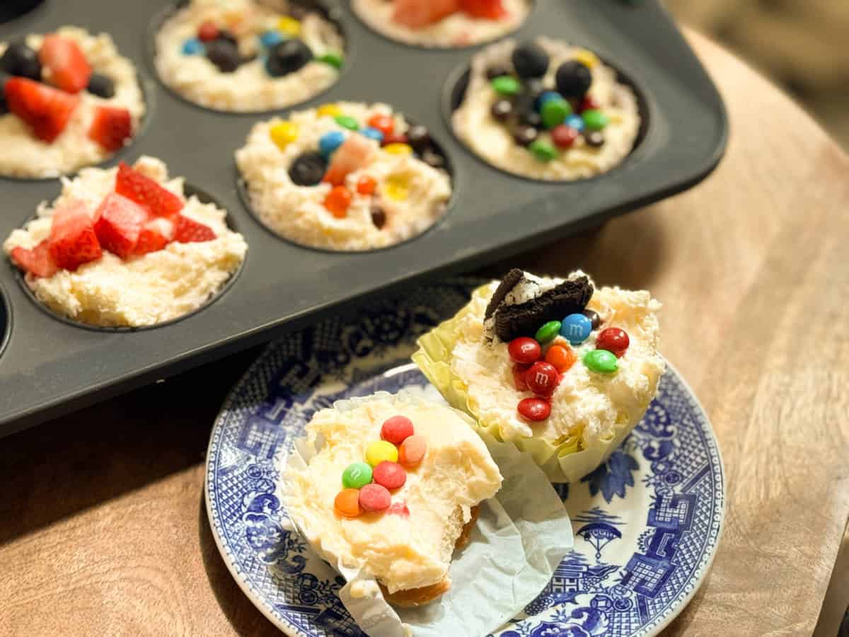 A muffin tin with partially prepared dessert cups topped with fruit and candy sits on a wooden surface. Two finished dessert cups with similar toppings are on a blue patterned plate in the foreground.