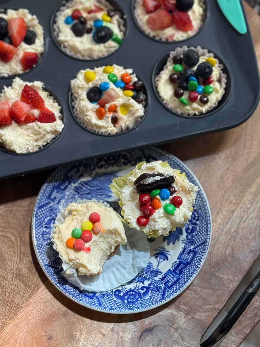A muffin tin with six cupcake liners filled with unbaked batter, topped with strawberries, blueberries, and chocolate candies sits on a wooden surface. Two decorated cupcakes rest on a blue and white plate nearby.