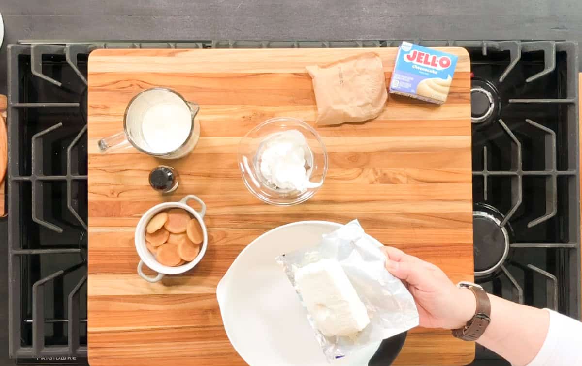A hand holds a block of cream cheese over a white plate on a wooden cutting board. Nearby are a bowl of whipped topping, a jug of milk, vanilla extract, vanilla wafer cookies, and a box of Jello pudding mix.