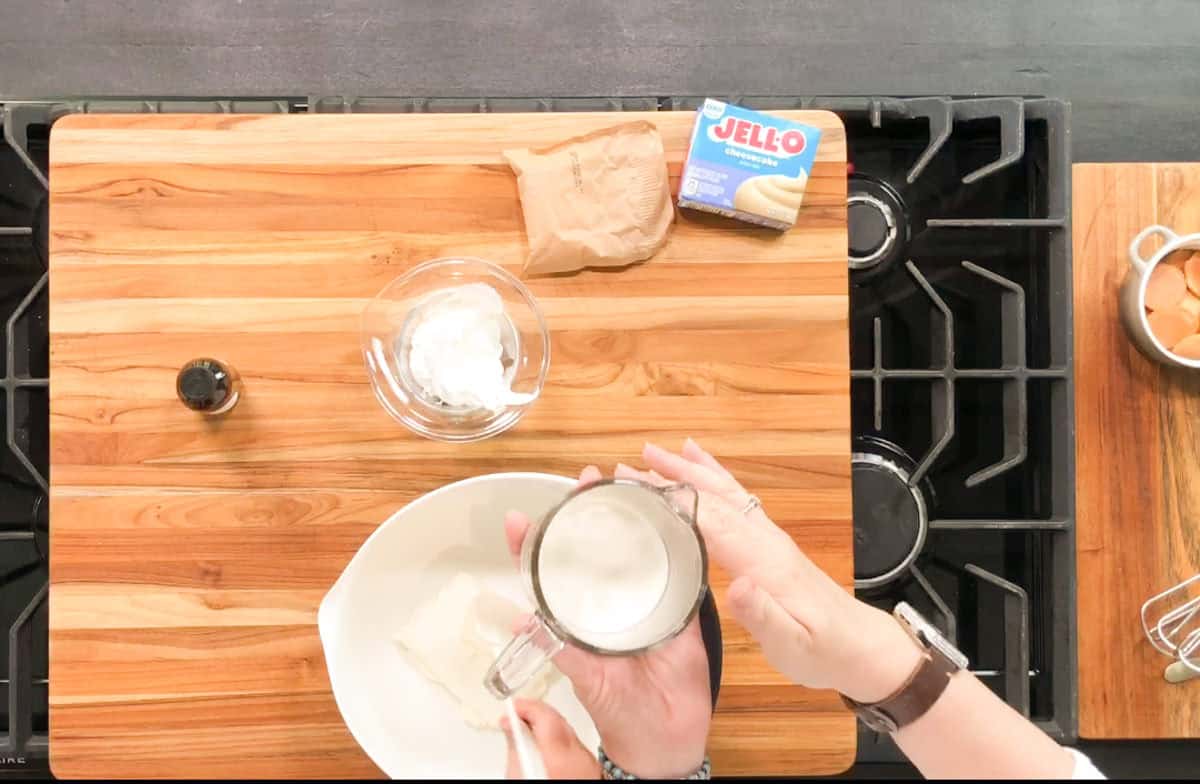 A person pours milk from a glass measuring cup into a white bowl with cream cheese. Other ingredients, including a vanilla extract bottle, whipped topping, instant pudding mix, and eggs, are on a wooden cutting board.