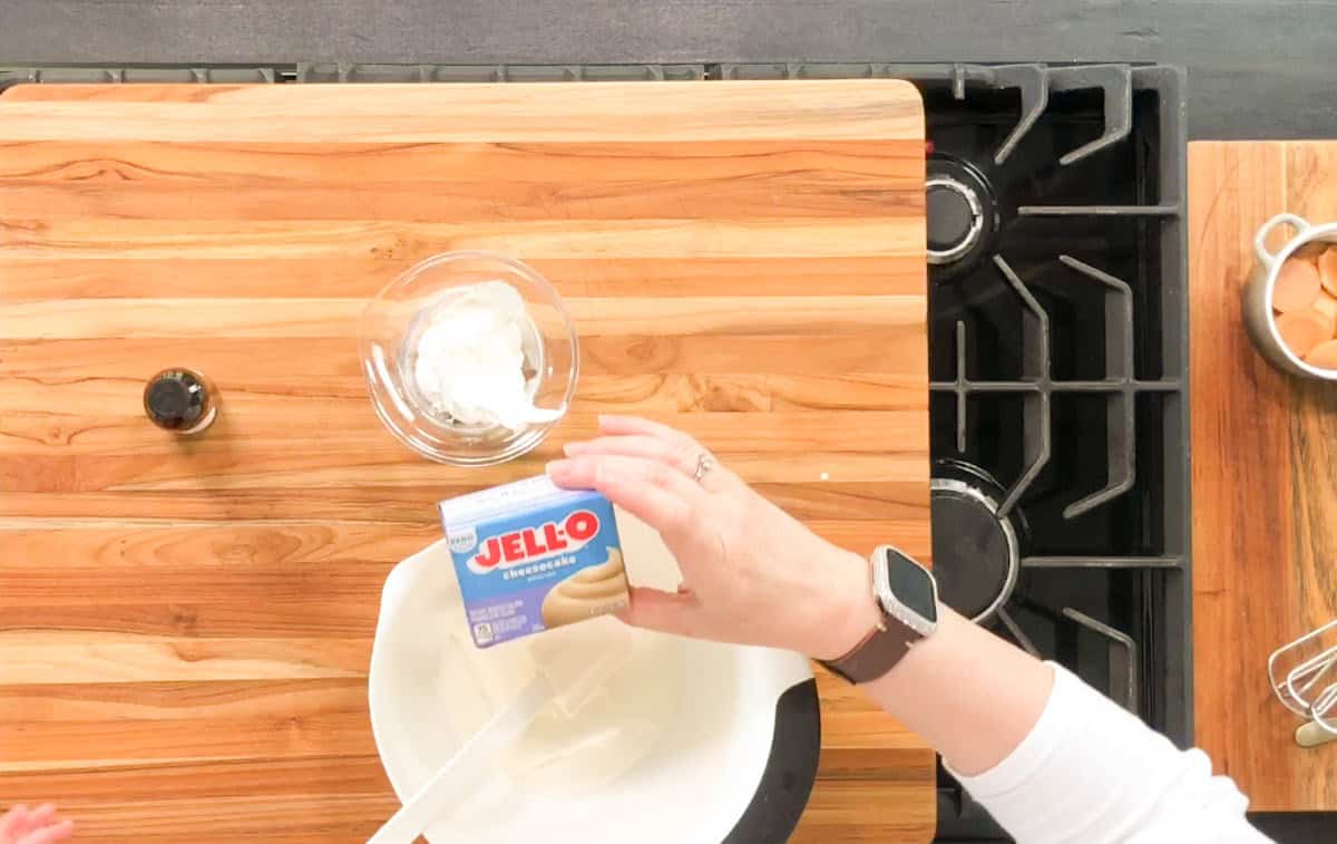 A person holds a box of Jell-O cheesecake instant pudding mix above a mixing bowl on a wooden countertop. Nearby are a small bowl of whipped topping, a bottle of vanilla, and a bowl of eggs next to a stovetop.
