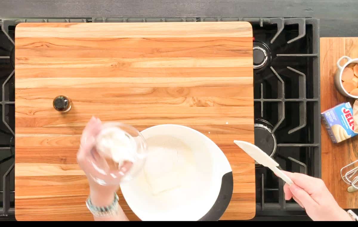 A person holds a spatula and a small bowl of cream cheese over a mixing bowl on a wooden cutting board, with vanilla extract, eggs, a mixer, and a box of Jell-O visible nearby on a stovetop.
