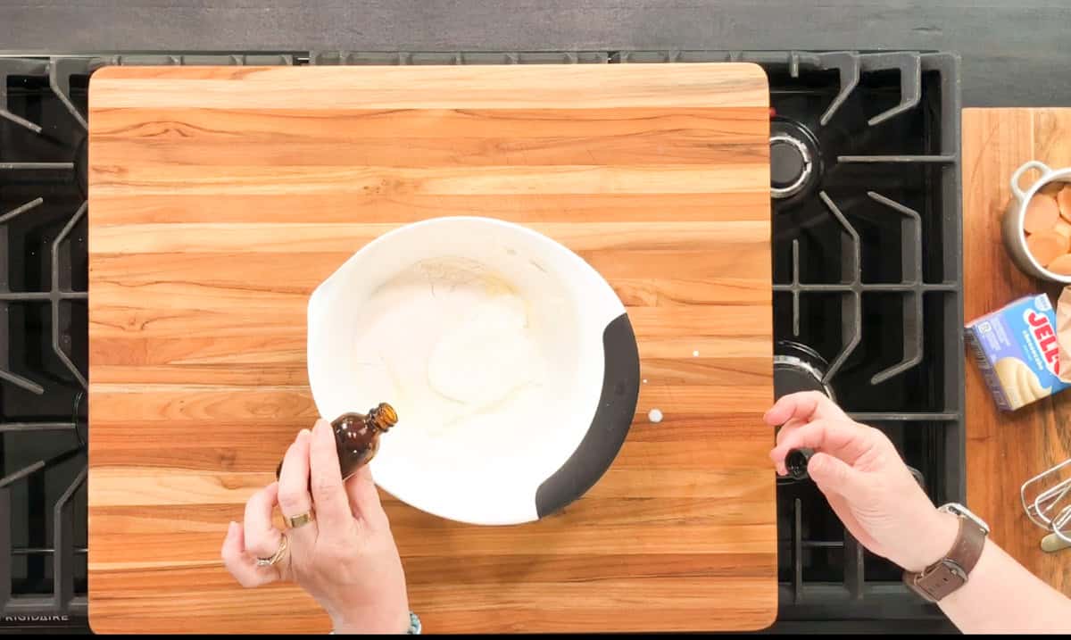 A person adds vanilla extract from a small bottle into a white mixing bowl with sugar on a wooden cutting board, with eggs and Jif peanut butter nearby on a kitchen counter.