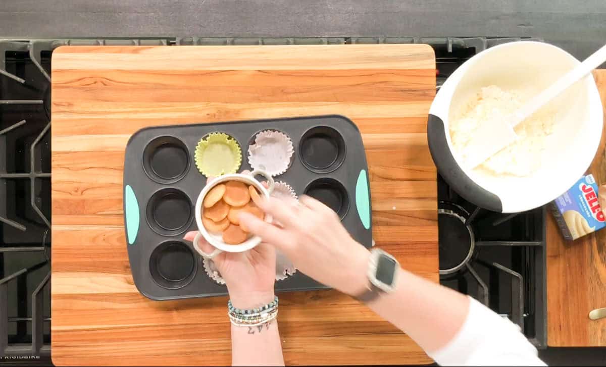 A person places sliced sausages into a muffin tin lined with paper cups. A bowl with a white spatula and a box of JELL-O are on a wooden cutting board nearby. The person wears a smartwatch and bracelets.
