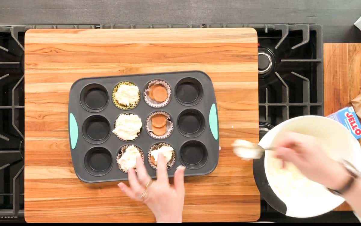A person is filling cupcake liners in a muffin tray with a creamy mixture using a spoon. The tray is on a wooden cutting board, and a white mixing bowl and a Jell-O box are nearby on a stovetop.
