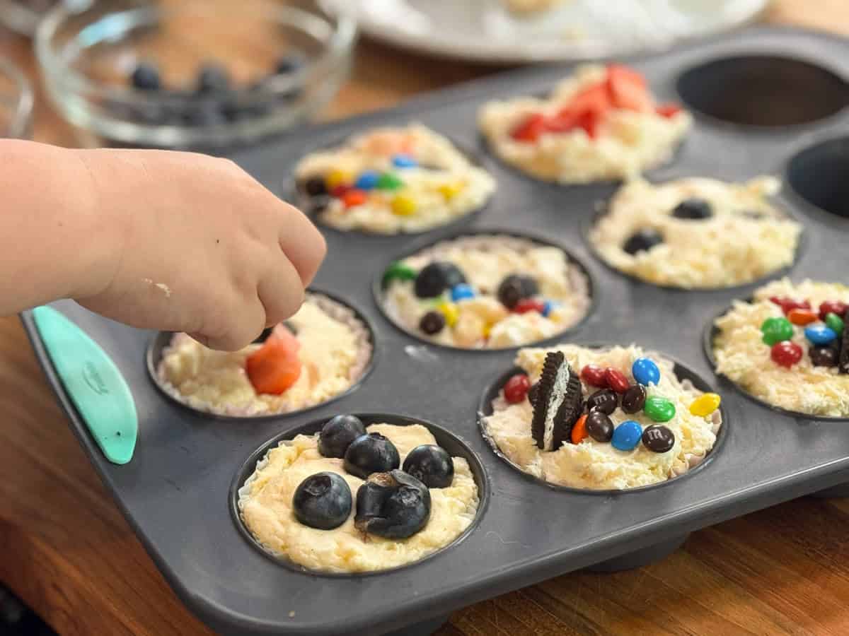 A child’s hand places a strawberry piece onto muffin batter in a muffin tin. Other muffin cups are topped with blueberries, mini chocolate candies, and cookie pieces. A bowl of blueberries is nearby on a wooden counter.