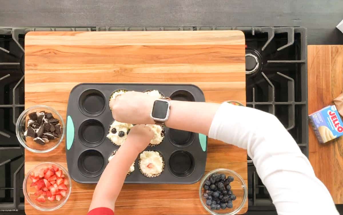 An adult and a child are decorating cupcakes in a muffin tin on a wooden cutting board. Bowls of chopped strawberries, blueberries, and crushed cookies surround them. A box of Jell-O is on the counter near the stove.