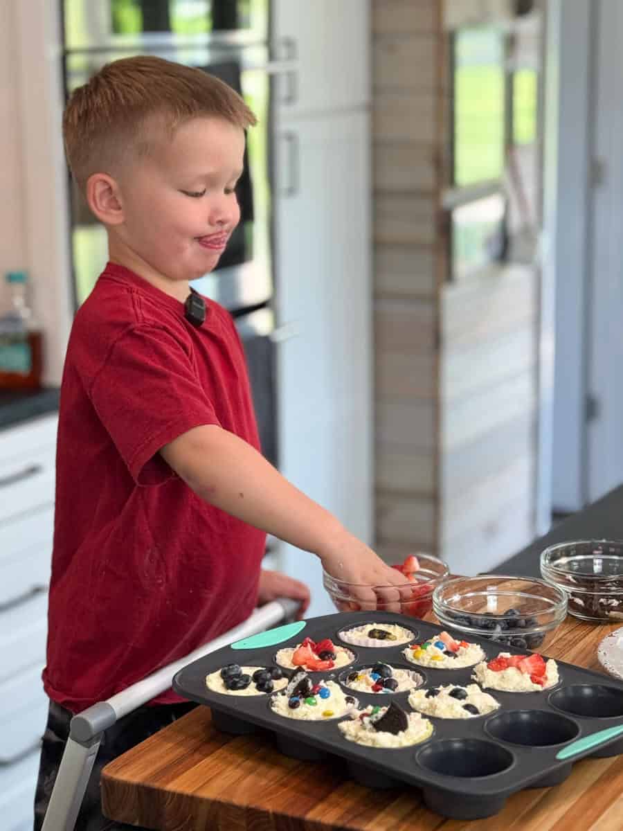 A young boy in a red shirt stands in a kitchen placing fruit into a muffin tin filled with batter. Several small bowls with toppings are on the counter next to him.