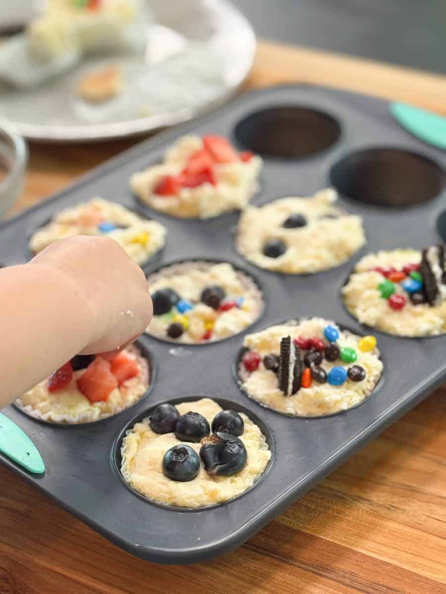 A hand places toppings onto rice-filled muffin cups in a muffin tray. Each cup is decorated with different items such as blueberries, strawberries, candy pieces, and cookie halves.