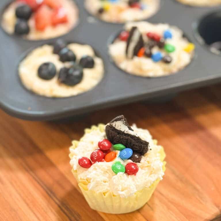 A muffin tin holds unbaked cupcake batter topped with strawberries, blueberries, Oreo pieces, and M&M candies. In the foreground, a single cupcake in a yellow wrapper is decorated with M&Ms and an Oreo piece.